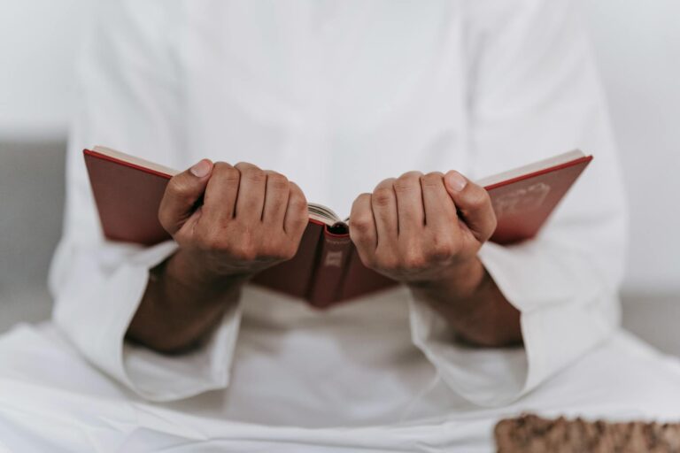 A man in traditional Islamic attire holds a Quran in a focused prayer session indoors.